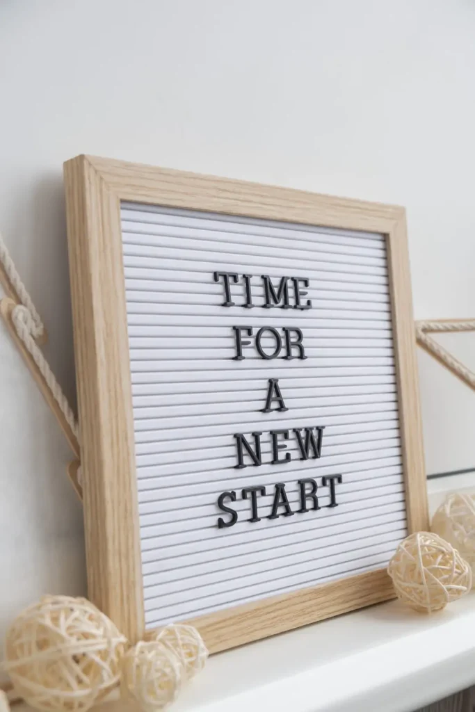 Auf einer Buchstabentafel mit Holzrahmen steht die Botschaft „ZEIT FÜR EINEN NEUANFANG“, umgeben von dekorativen Rattankugeln.