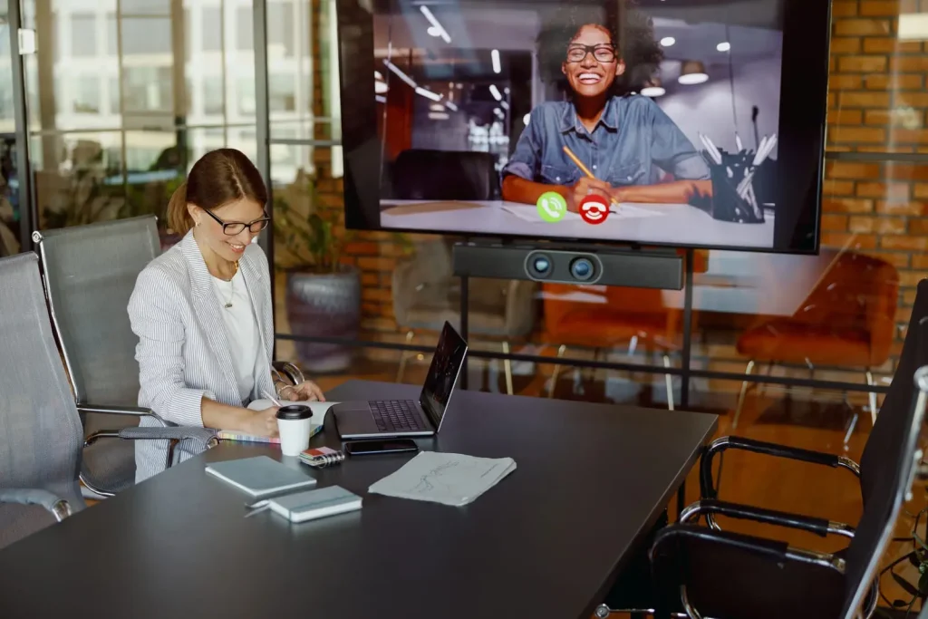 Eine Frau in einem Konferenzraum nimmt an einem Videoanruf auf einem großen Bildschirm teil.