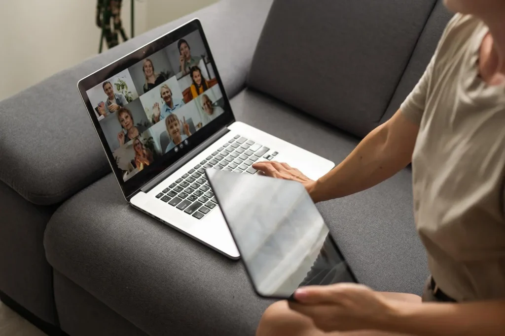 Person, die ein Tablet vor einen Laptop hält, auf dessen Bildschirm ein Videoanruf mit mehreren Personen angezeigt wird.